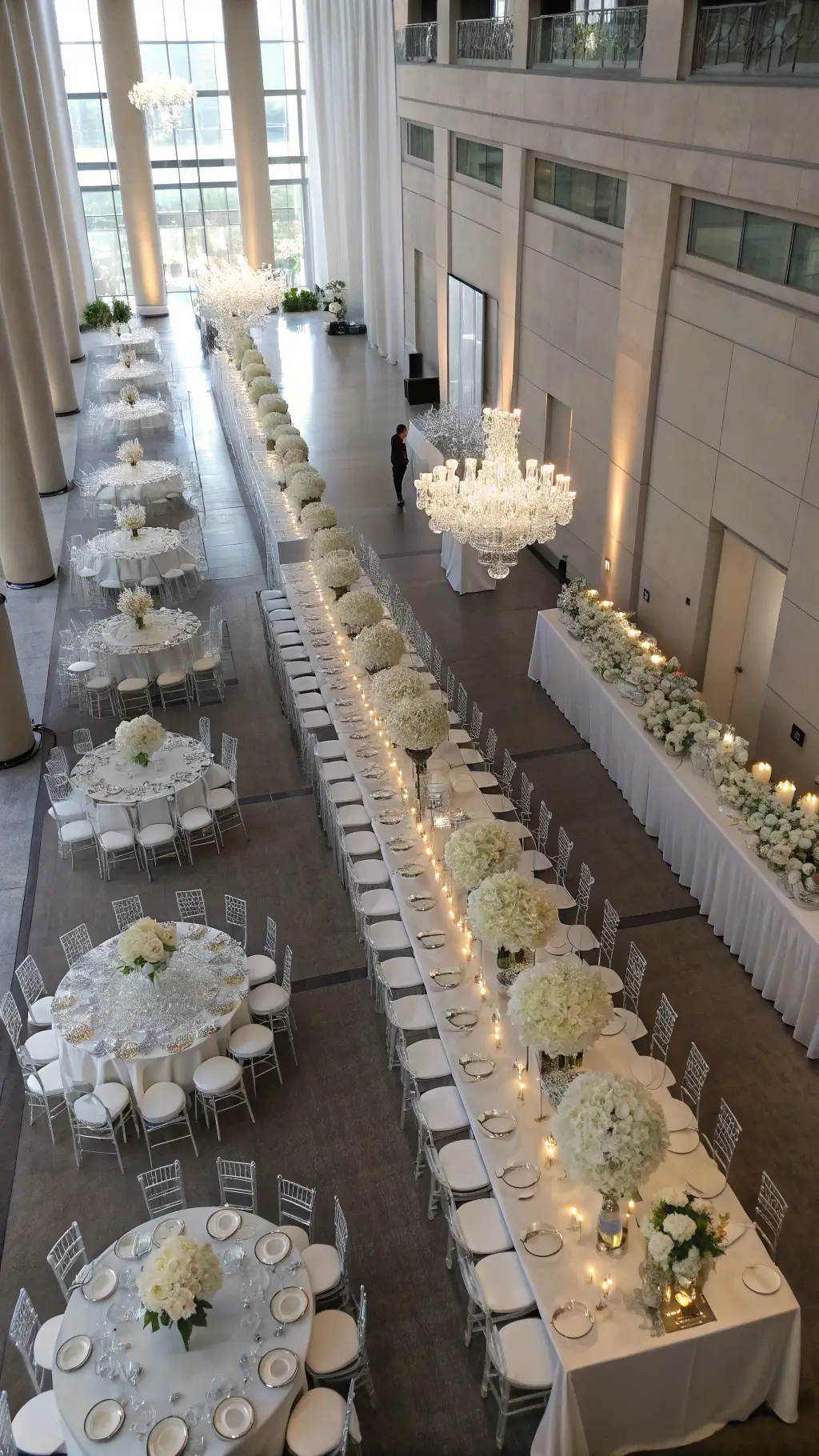 Creating White Christmas Wedding Centerpieces Aerial view of an elegant ceremony space with white-themed tablescapes; long and round tables feature white floral arrangements on clear risers, mercury glass votives, and crystal installations above; ghost chairs blend into the scene with natural light casting shadows on pewter flooring.