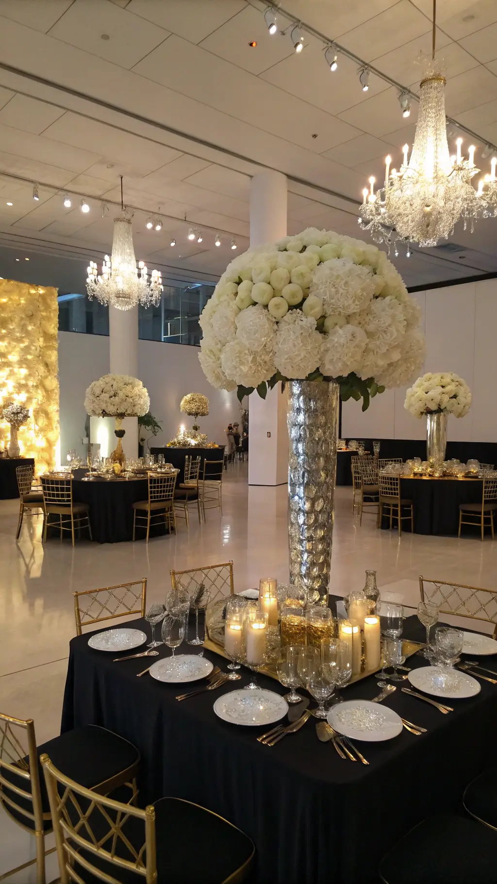 14 Wedding Floral Themes That Will Make Your Dream Day Come True (Number 6 is a Must-See!) A luxurious reception in a modern art gallery features mirrored tables with black silk linens, adorned with tall arrangements of white roses, dahlias, and orchids in gold geometric vessels. Crystal candelabras and votives enhance the dramatic lighting, while ghost chairs with gold cushions complement the metallic accents. A massive floral wall installation serves as the focal point, and the low angle shot captures chandelier reflections on mirrored surfaces, creating an infinity effect.