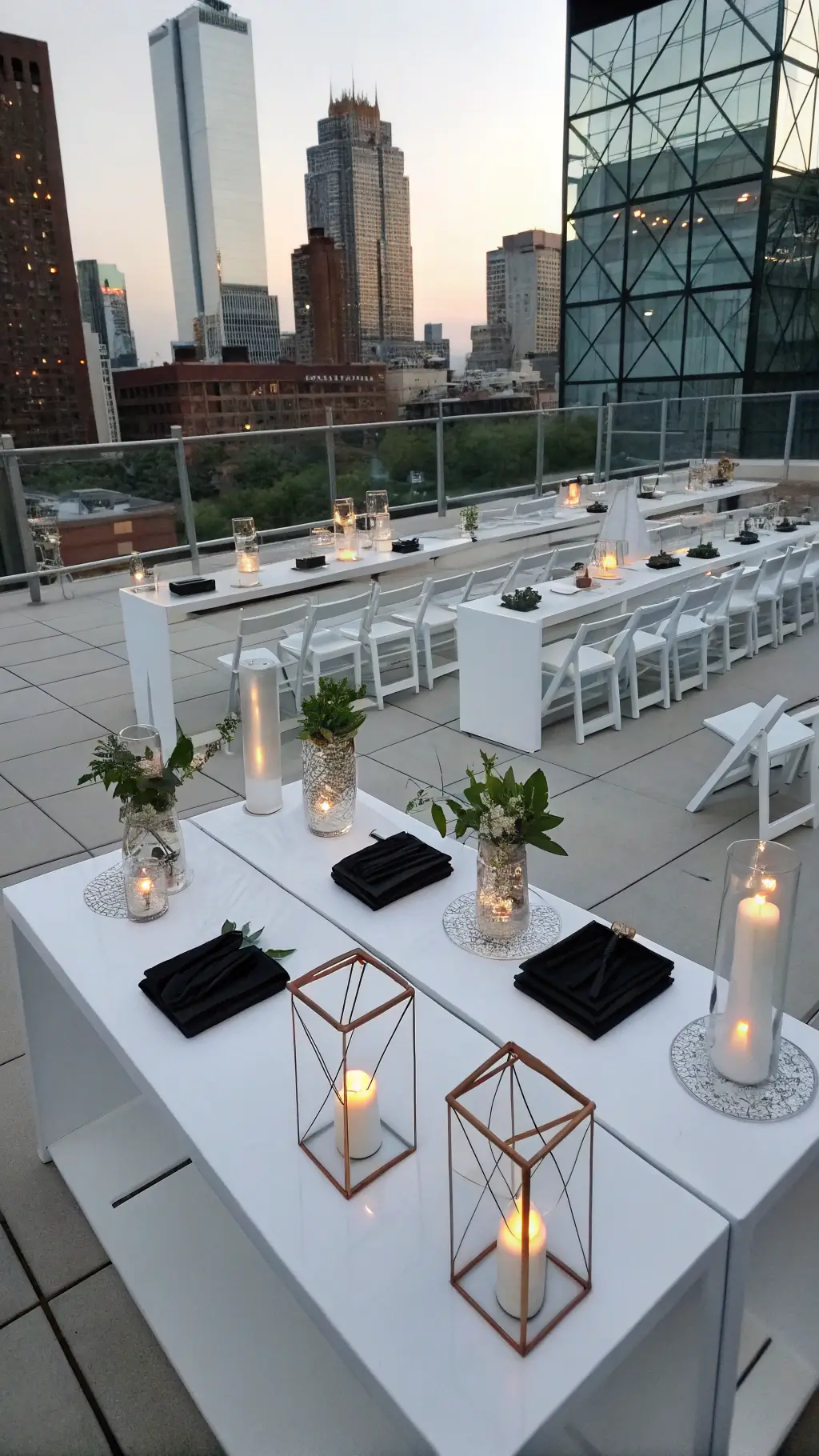 14 Wedding Floral Themes That Will Make Your Dream Day Come True (Number 6 is a Must-See!) A rooftop ceremony at blue hour featuring modern minimalist decor with white acrylic tables, black matte chargers, dramatic calla lily stems in tall vases, and geometric copper terrariums with candles. Ghost chairs surround the setup, with city skyline visible through glass walls, creating striking geometric patterns from a high-angle view.
