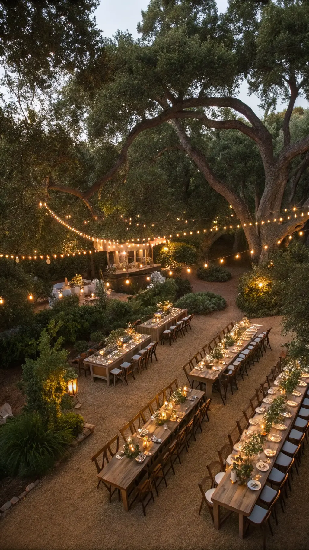 How to Create a Sophisticated Decor with Luxury Centerpieces? Aerial view of a garden wedding reception at twilight with U-shaped wooden tables under oak trees adorned with fairy lights. Tables are decorated with eucalyptus and rose garlands, brass vases, and votive candles. Cross-back chairs and vintage place settings complete the elegant scene, with market lights overhead twinkling like stars.
