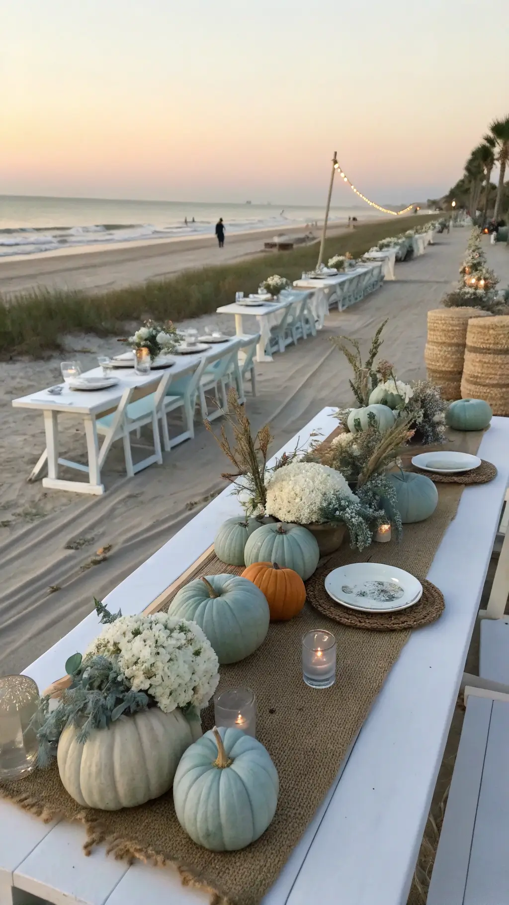 Elevate Your Table with a Stunning Pumpkin Centerpiece A scenic beachfront celebration at sunset showcasing coastal-themed pumpkin displays with whitewashed farm tables adorned with sea glass, driftwood, and blue-grey pumpkins, alongside white hydrangeas, dusty miller, and grasses; complemented by linen runners, rattan chargers, and shell accents, with the ocean horizon visible in the background.