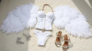How to Create a Victoria's Secret Angel Costume: Your Complete Guide A minimalist flatlay on plush white carpet featuring a complete Victoria's Secret Angel costume, including large white feathered wings, a white lace corset, garter belt, metallic strappy sandals, and crystal accessories, illuminated by natural skylight.
