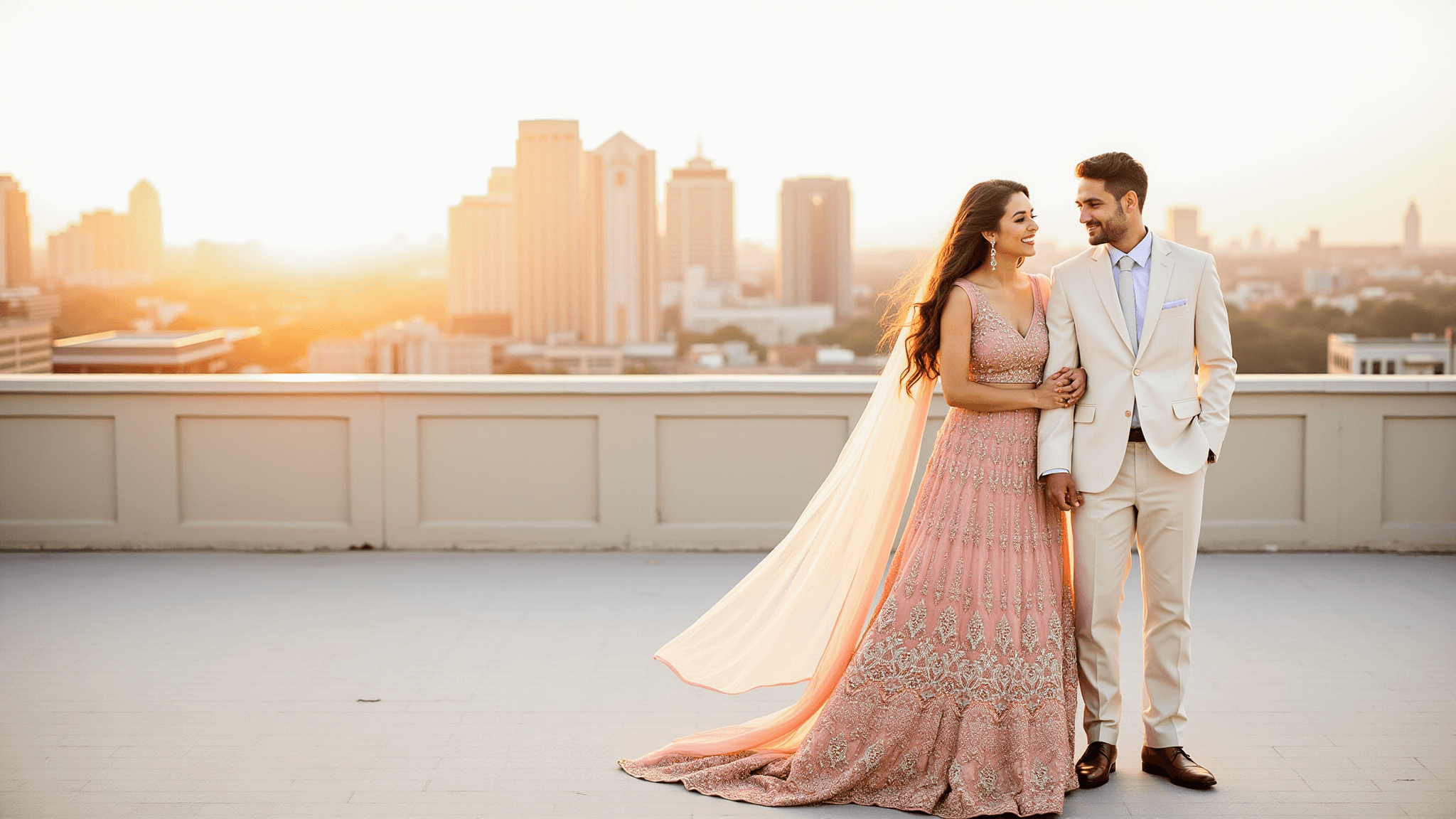 Couple on a rooftop at golden hour, woman in a blush pink lehenga with gold embroidery, man in an ivory suit, cityscape backdrop, warm sunset lighting, soft focus, editorial fashion aesthetic.